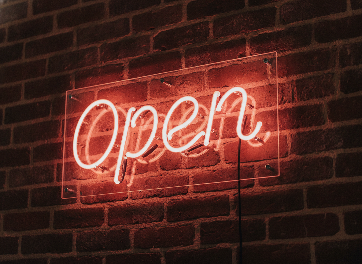 AI neon sign generator example — warm orange-pink cursive neon reading Open mounted on a dark exposed brick wall, rectangular acrylic backing plate with standoff screws and glow spilling onto the brickwork