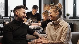 Create an 8-second cinematic advertising video.

Style:
Realistic modern European tattoo culture.
German tattoo studio aesthetic.
Clean, minimal, professional tattoo environment.
Neutral colors, warm low light, authentic atmosphere.
No American or flashy studio style.

Scene:
Inside a tattoo studio in Germany.
European tattoo artist working in the background.
A tattooed European man and a tattooed European woman sit nearby.
Natural body language, subtle eye contact, calm smiles.
Visible tattoos with realistic European tattoo styles.
Quiet, intimate moment, authentic connection.

Rules:
European people only.
German tattoo studio atmosphere.
No text.
No subtitles.
No logos.
No music.
No voice.
No exaggerated effects.