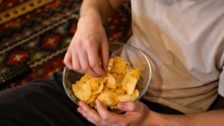 Create a realistic cinematic animation of the person naturally eating potato chips from the glass bowl. The person repeatedly reaches into the bowl, picks up one chip, brings it to their mouth, eats it, and returns their hand to the bowl in a smooth, natural loop. With every bite, the number of chips in the bowl gradually decreases, making the bowl visibly emptier over time. Ensure the chips disappear only when picked up and eaten, with realistic hand movements and natural timing. Keep the original camera angle, framing, clothing, lighting, background, and bowl position unchanged. Preserve realistic hand anatomy and chip textures. Static camera, no zoom, no sudden movements, no object deformation, no extra hands, and no changes to the person's appearance. Smooth, photorealistic motion throughout the animation.