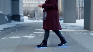 Elderly woman wearing a dark burgundy thermal-regulation coat with subtle glowing woven lines on the back and sides. 
Fitted dark slacks, refined black kinetic winter boots with soft blue luminescent soles. 
She carries a sleek transparent futuristic shopping basket with smooth edges. 
She is walking on the sidewalk of a futuristic city in the wintertime.