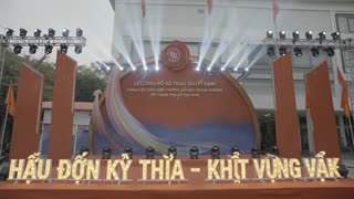 A cinematic event stage in front of a university building in Vietnam. The red ceremonial stage design with tall red panels and a large central emblem becomes animated. Soft sunlight, flags gently waving in the wind. The camera slowly pushes forward toward the stage. Dynamic light rays sweep across the structure. Golden particles and glowing lines appear. The central circular backdrop glows with blue and gold energy waves. Professional event atmosphere, dramatic lighting, high detail, realistic environment, cinematic motion, depth of field, 4K.