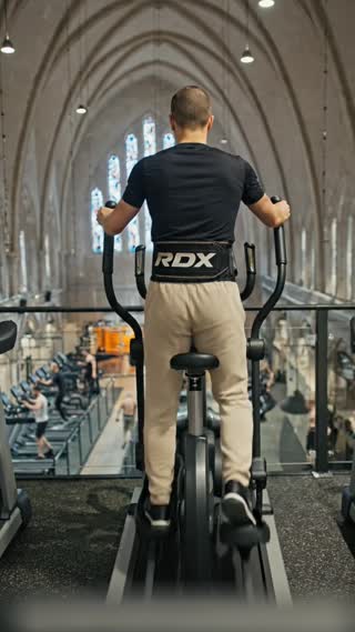 Créer une scène réaliste animée à partir de l’image fournie.
L’image doit être utilisée comme référence principale et rester strictement identique : même personnage, même position, même cadrage général, même salle de sport dans l’ancienne chapelle.

La caméra reste derrière la personne, qui est sur un vélo elliptique, et effectue un mouvement très léger et naturel, comme si un caméraman était debout derrière lui.
Le mouvement de caméra est lent, fluide et stable, avec une très légère oscillation cinématographique.

Le personnage pédale doucement sur le vélo elliptique, avec un mouvement naturel et régulier des bras et des jambes.

L’environnement doit rester fidèle à la salle Basic-Fit installée dans l’ancienne chapelle :

grandes fenêtres en vitraux gothiques visibles en arrière-plan

architecture de chapelle avec arches et hauteur sous plafond

salle de fitness en bas avec machines et équipements

En bas de la salle, quelques sportifs s’entraînent sur les machines (musculation, tapis de course, vélos), avec des mouvements naturels mais discrets.

Conserver un rendu extrêmement réaliste :

lumière naturelle venant des vitraux

ombres cohérentes

textures détaillées sur les vêtements

aucune transformation du personnage

Contraintes importantes :

utiliser exactement le même personnage et la même tenue que dans l’image

ne pas modifier la morphologie

ne pas modifier la couleur du jogging beige clair

ne pas modifier la ceinture RDX

ne jamais montrer le visage

pas d’effets artistiques, pas de style dessin, pas de style 3D

L’ambiance doit rester naturelle, stable et crédible, comme une vraie scène filmée dans la salle de sport installée dans cette chapelle. in a cinematic scene with professional lighting, shallow depth of field, and movie-quality visual effects