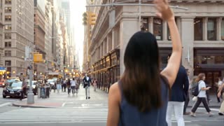 Generate a short, silent cinematic video set in a city with tall buildings, matching the provided reference images.
The scene contains no dialogue, no speech, no text, and no visible talking at any point; all storytelling is conveyed only through body language and movement.

A woman stands in the bottom-right foreground of the frame, facing away from the camera so that only the back of her head and upper torso are visible. She is standing on one side of a crosswalk, looking toward the opposite side of the street.

Across the crosswalk, near the buildings on the far side, her friend is visible walking along the sidewalk.

The woman raises her arm and waves enthusiastically and warmly, clearly trying to get her friend’s attention. The wave is friendly, energetic, and lasts for a brief moment.

Her friend does not notice her at all, does not look in her direction, and continues walking to the right, passing the building without stopping or reacting.

As the wave continues, the woman realizes she has not been seen. Her body language subtly shifts from excited and hopeful to quietly disappointed.

The friend continues walking and disappears completely out of sight.

The woman lowers her arm and remains standing, still facing the street as the moment passes.

The camera remains completely static, with no movement or reframing. The only motion in the scene is the woman’s wave and the friend walking away.

The clip ends with the woman still in place, her friend gone from view, and the missed connection clearly established — with silence throughout.
