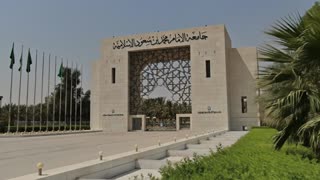 Photorealistic cinematic 4K video. The shot begins with an extremely smooth, slow-moving dolly camera outside the university gate shown in the reference image, focusing on architectural details and natural lighting as it gently approaches the entrance. The movement is fully stabilized and fluid with no shake or abrupt motion. The camera passes through the gate seamlessly with a soft cinematic flash transition into the interior scene.
The scene transitions into a spacious indoor hall inspired by the same architectural style of the building, featuring a modern, realistic design with a high ceiling and soft natural lighting. Inside the hall, four Saudi men wearing traditional Saudi attire appear in a balanced and professional composition at the center of the frame. In front of them is a medium-sized table with Arabic coffee cups and neatly arranged papers. One man holds an iPad and leads a focused professional discussion, suggesting a formal business or planning meeting.
The camera continues an ultra-smooth, slow cinematic glide through the space with shallow depth of field, soft lighting, realistic textures, and high-end commercial film quality, maintaining a calm, elegant, and highly professional visual tone throughout the scene.