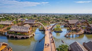 Using the provided image as the reference, create a cinematic architectural video sequence in 16:9. Faithfully preserve the project, its geometry, volumes, reefs, walkways, buildings, planted islands, and overall layout. Do not redesign the architecture or add any new elements.

In one continuous shot, begin with the wide aerial view. Move the camera smoothly over the water toward the heart of the project, following the main walkway and the curved shapes of the reefs. Lower the camera slightly to reveal the living spaces, waterways, and residents’ activities, then gradually rise and pull back to reveal the entire neighborhood and its relationship to the river. End on a broad, majestic panoramic view of the project.

Subtly animate the water, reflections, canoes, vegetation, and people. Keep natural daylight, a believable tropical atmosphere, and restrained colors. The camera movement should be smooth, sweeping, and spectacular, like footage captured by a professional drone. Realistic architectural visuals, with a clear sense of depth and scale.

No design changes, no building distortions, no additional structures, no abrupt movements, no text, no logos, and no artificial transitions. in a cinematic scene with professional lighting, shallow depth of field, and movie-quality visual effects