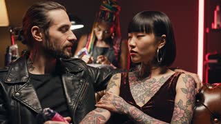 Create an 8-second cinematic advertising video.

Style:
Modern, edgy, tattoo culture aesthetic.
Dark tattoo studio environment, warm low light, subtle neon accents.
Cinematic slow motion, shallow depth of field.

Scene:
Inside a tattoo studio.
A tattoo artist works in the background.
In the foreground, a tattooed man and a tattooed woman sit nearby.
They exchange a look, smile subtly.
Their tattoos are visible and feel personal.
Quiet, intimate chemistry, authentic moment.

Rules:
No text.
No subtitles.
No logos.
No music.
No voice.
Stylized but realistic atmosphere.
