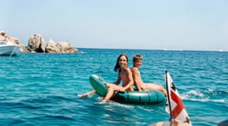 Create a short, realistic video animation from this image. Preserve the exact identity, facial features, and proportions of the people — no alterations or stylization. Maintain full realism.

Animate the ocean water with natural, gentle motion: soft waves, subtle ripples, and realistic light reflections moving across the surface. The inflatable float should move slightly with the water in a smooth, buoyant way.

Add small, natural human movements: slight body balance adjustments, minimal head motion, and subtle breathing — no exaggerated or artificial gestures. Hair should move lightly with the breeze.

Include a soft sea breeze effect: gentle movement in the water and a barely noticeable shift in the surrounding environment. The distant background (rocks, boat, horizon) should remain stable with only minimal atmospheric motion.

Lighting should stay consistent with a sunny day, with natural highlights shimmering on the water. Colors must remain realistic and balanced, not oversaturated.

Motion must be smooth and cinematic — no abrupt transitions, no jitter, no distortion. Keep the camera stable (no zoom or shake), as if filmed from a fixed position.

Loop seamlessly.
