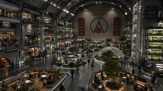 Live-action photorealistic wide interior shot of a massive multi-level atrium inside a futuristic Mars settlement habitat, an immense arched vaulted ceiling with exposed structural trusses and warm overhead lighting, the far wall a smooth solid structure with no signage or text of any kind. Multiple stacked levels of interior balconies line the left side, warmly lit interior offices and gathering spaces visible through open facades, no labeling anywhere. On the right, a tall vertical farming structure holds stacked rows of illuminated leafy greens under grow lights. The ground floor centers on a large glass geodesic dome housing a full-grown tree, surrounded by open plazas, smaller garden domes, and sunken communal seating areas with wooden benches and warm under-lighting where small groups of people sit and talk. Dozens of people in casual Mars colony clothing — fitted jumpsuits, layered utility jackets, practical footwear — walk across the open central floor between the plazas. The room is fully enclosed with no windows or openings to the outside, all illumination coming from engineered ceiling and ambient lighting. Camera holds a high wide static angle looking down across the entire atrium, with a slow, almost imperceptible push-in. Shot on real film camera aesthetic with natural lens grain, warm and cool lighting contrast, documentary-style realism, no CGI or animated look, bustling yet orderly atmosphere emphasizing a fully self-contained, thriving society.