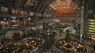 Live-action photorealistic wide interior shot of a massive multi-level atrium inside a futuristic space habitat, an immense vaulted ceiling with exposed structural trusses and industrial framework arching overhead, the far wall now fully enclosed with solid paneling and structural framework in place of the arched window — no view to the outside world, all illumination coming from engineered overhead and ambient lighting rather than any exterior light source. Multiple stacked levels of interior balconies and walkways line both sides of the space, lit storefronts and offices visible along the upper floors, one clearly marked "ARES HABITAT" with a level marker "02" beside it, and another labeled "03 AGRICULTURE" near a tall vertical farming structure with illuminated stacked plant racks growing rows of greenery. In the center of the ground floor, a large glass-domed greenhouse structure houses a full-grown tree surrounded by an open communal seating area with round tables and warm under-lighting, people seated and conversing. To the lower left, another sunken lounge area with tables, benches, and potted plants is filled with seated people. Scattered individuals in casual and utility-style clothing walk across the open central floor between the seating areas, some near a rounded doorway entrance on the lower left leading to a warmly lit interior room. The architecture combines exposed industrial metal framework with warm wood-toned seating areas and cool structural surfaces, soft ambient down-lighting throughout, camera holding a high wide static angle looking down across the entire atrium, shot on real film camera aesthetic with natural lens grain, documentary-style realism, no CGI or animated look, bustling yet orderly atmosphere emphasizing a fully self-contained.