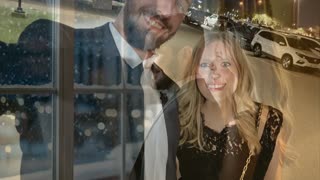 A couple stands close together in a cozy, softly lit room at night, watching snow fall gently outside the window. They share a warm, affectionate smile. The scene then transitions to the same couple standing together in front of the Bellagio Hotel in Las Vegas at night. Snow is falling lightly around them, and the iconic Bellagio fountains are erupting in a spectacular, illuminated water show behind them. Cinematic style, warm and cool lighting contrast, shallow depth of field, smooth transition.