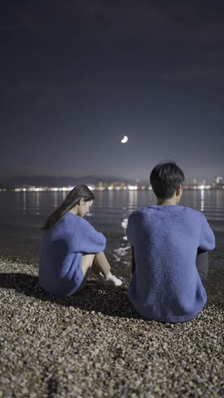 The same nighttime lakeside scene continues with the Korean couple sitting far apart in silence. The camera stops its subtle movement and becomes completely still, holding a wide vertical 9:16 composition with large negative space above and between them.

Both remain in the same positions — the woman slightly curled inward, the man facing outward — with no interaction or eye contact. Their distance remains unchanged.

The environment grows slightly quieter and darker, with a gentle dimming of ambient light. The city reflections on the water soften, and the ripples become calmer, almost still. The sky appears deeper and more muted, with fewer visible details.

Very minimal motion: faint wind in the hair and clothing, barely perceptible. The overall feeling is stillness and quiet acceptance.

Lighting remains natural, uneven, and slightly grainy, like a real low-light camera. No dramatic highlights, no cinematic effects.

The focus is on silence, space, and the feeling that something remains without needing to be resolved.