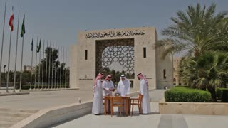 Photorealistic cinematic 4K video. The shot begins with a slow forward camera movement approaching the university gate shown in the image, with smooth, natural motion creating a realistic cinematic feel. As the camera gets closer to the entrance, four Saudi men wearing traditional attire (thobes and ghutras) are revealed standing in the center.

They are positioned around a medium-sized table placed in harmony with the entrance layout. On the table are coffee cups and neatly arranged papers. One man is holding an iPad, interacting with the others as if presenting or explaining something, while the rest are attentively focused on him in a professional discussion stance.

The setting is an outdoor courtyard with clean flooring, palm trees, and the architectural gate clearly visible in the background. Natural daylight with realistic shadows. The camera maintains subtle motion, focusing on composition and the interaction between the subjects.

No text, no dialogue, no music. Only soft natural ambient sound.