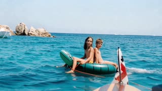 Create a short, realistic video animation from this image. Preserve the exact identity, facial features, body proportions, and original facial expressions of the subjects. The children must not smile, laugh, or change expression — their faces should remain exactly as in the original photo.

Maintain full realism: no stylization, no beautification, no artificial changes.

Animate the ocean water with natural, gentle motion: soft waves, subtle ripples, and realistic sunlight reflections moving across the surface. The inflatable float should drift slightly and naturally with the water, following realistic buoyancy.

Human movement must be minimal and physically accurate: only subtle balance adjustments caused by the water, very slight posture shifts, and natural micro-movements (e.g., breathing). No added emotions, gestures, or interactions.

Hair may move slightly with a soft sea breeze. The environment (rocks, distant boat, horizon) should remain stable, with only minimal atmospheric motion.

Lighting must remain consistent with the original image: bright daylight, natural highlights on the water, realistic color tones (no oversaturation).

Motion must be smooth and cinematic: no abrupt movement, no jitter, no morphing, no distortion. Camera must remain fixed and stable (no zoom, no panning).