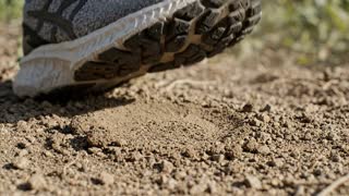 Close-up shot, live-action photorealistic, a sneaker stepping down into soft outdoor dirt, the shoe's sole pressing into the loose earth and leaving a clear textured impression, small clumps of soil shifting and scattering slightly around the point of contact, natural daylight casting a soft shadow beneath the shoe, fine dust or dry soil particles kicked up faintly on impact, shallow depth of field keeping the shoe and ground in sharp focus while the background falls into a soft natural blur, shot on real film camera aesthetic with natural lens grain, true-to-life color grade, slow motion capturing the moment of contact, documentary-style realism, no CGI or animated look, grounded and tactile mood