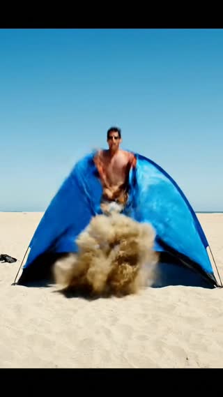 A single white Argentine man in his late 30s, with short dark hair and a relaxed expression, wearing casual beach shorts and sunglasses, appears enjoying himself on a sunny beach, reclining on a lounge chair while reading a paperback book. He then gets up and begins assembling a large, blue camping tent on the sand. After successfully finishing the assembly, he prepares to enter the tent for an afternoon nap. Suddenly, a powerful gust of wind blows, filling the interior of the tent completely with fine, dry sand. The man, now frustrated, puts on a bright yellow construction helmet and a worker's high-visibility vest. He grabs a metal shovel and begins vigorously shoveling the sand out from inside the tent as if it were construction material, treating the tent like a worksite. Beads of sweat form on his forehead under the hot sun; he pauses to wipe his brow with the back of his arm, shaking his head in disbelief and exasperation, as if muttering "you've got to be kidding me" at the absurdity of the forced labor.

Cinematic wide shot establishing the beach scene, cutting to medium shots of the assembly and close-ups of his frustrated expressions. Handheld camera movement conveys the sudden chaos of the wind. Bright, high-noon sunlight with harsh shadows creates a hot, exhausting atmosphere. Ambient sound of waves, wind gusts, and the sound of shoveling sand.