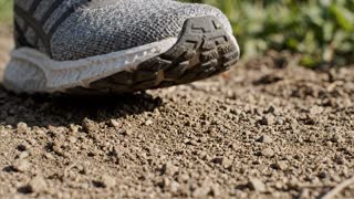 Close-up shot, live-action photorealistic, a sneaker stepping down into soft outdoor dirt, the shoe's sole pressing into the loose earth and leaving a clear textured impression, small clumps of soil shifting and scattering slightly around the point of contact, natural daylight casting a soft shadow beneath the shoe, fine dust or dry soil particles kicked up faintly on impact, shallow depth of field keeping the shoe and ground in sharp focus while the background falls into a soft natural blur, shot on real film camera aesthetic with natural lens grain, true-to-life color grade, slow motion capturing the moment of contact, documentary-style realism, no CGI or animated look, grounded and tactile mood