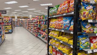 Animate this image with a very subtle, realistic cinematic camera movement. Apply a slow, smooth forward dolly-in with a gentle sideways camera drift, creating natural depth and parallax between the foreground candy shelves and the background. Keep the movement minimal, steady, and physically realistic, as if filmed by a professional camera operator inside a real supermarket. Preserve all original product packaging, colors, text, shelf positions, lighting, and proportions exactly as they are. No object movement, no floating products, no morphing, no flickering, no warping, no artificial zoom, and no sudden camera movements. Maintain realistic indoor lighting and natural reflections. One continuous shot, approximately 5–7 seconds, photorealistic, stable, and seamless.