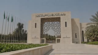 Photorealistic cinematic 4K video. The shot begins with an extremely smooth, slow, stabilized dolly movement outside the university gate shown in the reference image, emphasizing architectural details and soft natural lighting as the camera gently approaches the entrance. The motion is fluid, continuous, and perfectly stabilized with no shake or abrupt cuts, creating a calm cinematic tone.

The camera passes through the gate seamlessly with a soft cinematic transition into the interior. The scene reveals a spacious indoor hall inspired by the same building, featuring modern Islamic architectural design with elegant arches, geometric patterns, and subtle traditional detailing, while maintaining a realistic contemporary style with a high ceiling and soft natural lighting.

Inside the hall, four Saudi men wearing traditional Saudi attire (thobe and red shemagh only, no white ghutra) appear in a balanced and professional composition at the center of the frame. In front of them is a medium-sized table with Arabic coffee cups and neatly arranged papers. One man holds an iPad and leads a focused professional discussion, suggesting a formal business or planning meeting.

The camera continues an ultra-smooth, slow cinematic glide through the space with shallow depth of field, soft natural lighting, realistic textures, and high-end commercial film quality, maintaining a calm, elegant, and professional visual atmosphere throughout the scene.