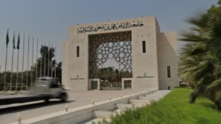 Photorealistic cinematic 4K video. The shot begins with a slow, stabilized dolly movement outside the university gate shown in the reference image, focusing on architectural details and natural lighting as the camera approaches the entrance. The camera smoothly passes through the gate, transitioning seamlessly into a spacious indoor area inspired by the same architectural design of the building. The space is a large, open, high-ceiling hall with a modern and realistic architectural style.

Inside this wide hall, four Saudi men wearing traditional Saudi attire appear in the center of the frame in a balanced and professional composition. In front of them is a medium-sized table with Arabic coffee cups and neatly arranged papers. One man is holding an iPad and leading a focused professional discussion with the group, suggesting a formal planning or business meeting.

The camera continues a smooth cinematic glide through the space, maintaining realistic lighting, shallow depth of field, natural motion blur, and high-end commercial production quality.