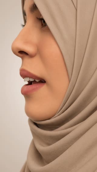 Buat animasi edukasi 3D vertikal format 9:16 dari gambar seorang muslimah berhijab yang sedang memperagakan makhraj huruf **ظ**. Pertahankan wajah, jilbab, pencahayaan, sudut kamera, dan gaya visual realistis 3D seperti gambar asli.

Animasi dimulai dari posisi mulut sedikit terbuka. Gerakkan ujung lidah secara perlahan ke arah tepi bawah gigi seri atas, lalu buat ujung lidah sedikit keluar di antara gigi atas dan bawah. Posisi lidah harus terlihat jelas menyentuh atau sangat dekat dengan ujung gigi seri atas, bukan berada di belakang gigi.

Saat mengucapkan huruf **ظ**, buat pangkal lidah sedikit terangkat sehingga menghasilkan karakter suara tebal. Bibir tetap rileks dan tidak terlalu monyong. Rahang terbuka secukupnya. Gerakan mulut harus natural, lembut, dan anatomis.

Fokus kamera tetap pada gerakan lidah, gigi seri atas, dan bentuk bibir. Jangan mengubah posisi kepala. Jangan menambahkan teks, subtitle, simbol, panah, logo, atau elemen grafis lain. Gunakan latar belakang polos, gerakan halus, detail anatomi yang jelas, dan hasil akhir seperti video pembelajaran tajwid profesional.


