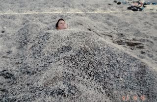 Create a short, highly realistic video animation from this photograph. Preserve the exact identity, facial features, proportions, and original facial expression of the child. The expression must remain unchanged — calm and relaxed, as in the original image. No added emotions, no smiling changes, no exaggeration.

Maintain full realism: no stylization, no beautification, no artificial smoothing.

The child is buried under sand and enjoying a peaceful moment — therefore, movement must be extremely minimal. Animate only very subtle breathing, visible as barely perceptible, natural micro-shifts in the sand around the chest area. The body must remain otherwise completely still.

The sand should feel real and natural, with only very slight surface settling — no visible sliding, no dramatic particle motion, no displacement that breaks the stillness.

No movement from nearby objects (such as towels, stones, or background elements). The environment should feel quiet and stable, almost frozen in time.

Lighting must remain consistent with the original: soft daylight, natural shadows, slightly muted tones. No color exaggeration.

Motion must be smooth and nearly imperceptible: no sudden changes, no jitter, no morphing, no distortion. Camera must remain completely fixed.

Overall feeling: serene, warm, and still — like a real moment gently coming to life without disturbing its calmness.