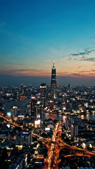 A breathtaking cinematic aerial shot of Bangkok, Thailand, at blue hour transitioning into night. Skyscrapers, busy roads, illuminated buildings, and an expanding modern city, symbolizing rising electricity demand. Smooth forward drone movement over the skyline, realistic Thai urban architecture, rich teal shadows and warm orange city lights, subtle atmospheric haze, dramatic cinematic lighting, photorealistic, professional energy documentary, vertical 9:16. No text, no subtitles, no watermarks.