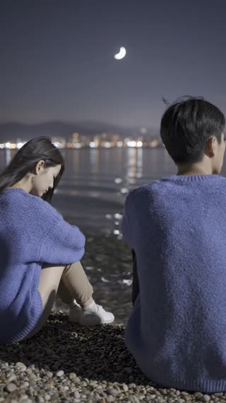 A quiet nighttime lakeside scene continues with the same Korean couple sitting far apart. The composition remains wide in a vertical 9:16 frame, but the camera now begins a very slow, subtle push-in toward the space between them, not focusing on either person directly.

The woman on the left remains still, head lowered; the man on the right continues to look outward. There is no interaction or eye contact. The emotional distance remains unchanged.

Very subtle environmental motion: gentle water ripples reflecting distant city lights, faint wind moving hair strands and soft fabric slightly. The atmosphere feels slightly heavier than before, with a marginally darker tone and cooler color temperature.

Lighting remains natural and imperfect, with uneven shadows. Slight grain and low-light texture present, like a real camera capturing a quiet night scene.

The focus is on the empty space between them, emphasizing separation and quiet tension.

no fast zoom, no close-up faces, no dramatic gestures, no turning heads, no interaction, no emotional exaggeration, no cinematic lens flare, no bright lighting, no perfect symmetry

