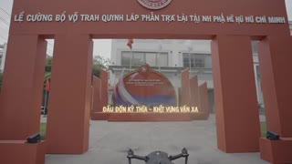 FPV drone flying toward a ceremonial red event stage set in front of a university building in Vietnam. The drone flies smoothly through tall red panels like a gateway and approaches the glowing central stage. Flags waving, dynamic sunlight, particles and light streaks appearing around the structure. The slogan “Dấu ấn kế thừa – Khát vọng vươn xa” lights up dramatically. Cinematic lighting, epic atmosphere, realistic motion, depth of field, 4K, smooth FPV movement.