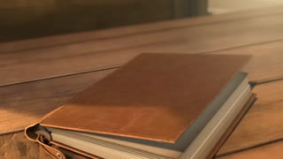 A vintage leather-bound photo album rests on a rustic wooden table, its pages slowly turning with a gentle, magical motion. Each spread features two warm, nostalgic photographs: one showing a smiling young couple holding hands as they approach their new home's front door under soft afternoon light, the next displaying a multi-generational family gathered on a porch with flowering window boxes, laughing together. The lighting is warm and golden, casting soft shadows across the album's textured pages. A whimsical, uplifting piano melody plays softly in the background. A warm, gentle voiceover says, "Through every story, we are thankful to help write the next chapter." On the final page, elegant script reads "Geoff and Krista Jones are your real estate experts," with stylized contact details printed discreetly at the bottom. Cinematic shallow depth of focus, intimate close-up shots of turning pages, warm nostalgic atmosphere